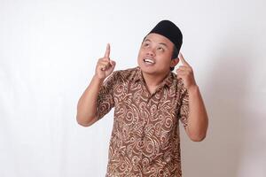 Portrait of excited Asian man wearing batik shirt and songkok smiling and looking at the camera pointing with two hands and fingers to up. Isolated image on gray background photo