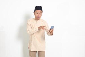 Portrait of surprised Asian muslim man in koko shirt with skullcap holding mobile phone, showing shocked face expression. Advertising and social media concept. Isolated image on white background photo