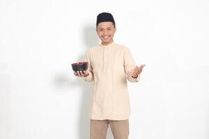 Portrait of excited Asian muslim man in koko shirt with skullcap showing and holding an empty plate. Bowl template for food brand. Isolated on white background photo