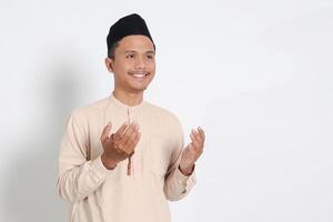 Portrait of religious Asian muslim man in koko shirt with skullcap praying earnestly with his hands raised, holding islamic beads. Devout faith concept. Isolated image on white background photo