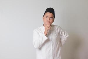 Portrait of Asian muslim man in white koko shirt with skullcap thinking about question with hand on chin. Isolated image on white background photo