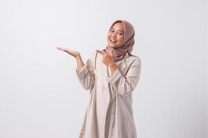 Portrait of excited Asian hijab woman in casual suit pointing and showing product in her side with finger. Businesswoman concept. Isolated image on white background photo