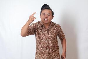 Portrait of unpleasant Asian man wearing batik shirt and songkok showing cynical unhappy angry facial expression pointing forward, giving warn. Isolated image on gray background photo