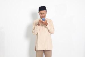 Portrait of young excited Asian muslim man in koko shirt with peci holding mobile phone with smiling expression on face. Social media and internet concept. Isolated image on white background photo