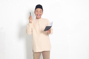 Portrait of attractive Asian muslim man in koko shirt with peci reading a book, telling that he has an idea while pointing finger and pen. Isolated image on white background photo