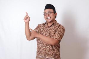 Portrait of excited Asian man wearing batik shirt and songkok smiling and looking at the camera pointing with two hands and fingers to the side. Isolated image on gray background photo