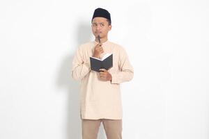 Portrait of confused Asian muslim man in koko shirt with peci reading a book, thinking about an idea with hand holding pen on chin, looking away. Isolated image on white background photo