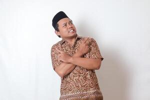 Portrait of confused Asian man wearing batik shirt and songkok with crossed hands, pointing sideways, making choice, choosing between two objects. Isolated image on gray background photo