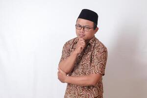 Portrait of confused Asian man wearing batik shirt and songkok standing against gray background, thinking about question with hand on chin photo
