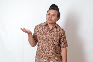 Portrait of confused Asian man wearing batik shirt and songkok showing empty hand. Isolated image on gray background photo