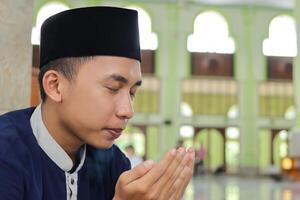 Portrait of religious Asian man in muslim shirt and black cap praying with hand raised inside the public mosque photo