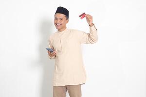 retrato de atractivo asiático musulmán hombre en koko camisa con casquete participación un móvil teléfono y presentación crédito tarjeta. aislado imagen en blanco antecedentes foto