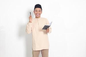 Portrait of attractive Asian muslim man in koko shirt with peci reading a book, telling that he has an idea while pointing finger and pen. Isolated image on white background photo