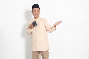 Portrait of attractive Asian muslim man in koko shirt with peci holding a mug, drinking water, quenching thirst after fasting. Isolated image on white background photo