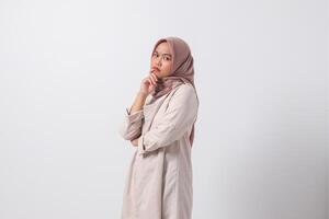 Portrait of confused Asian hijab woman in casual suit thinking with hand on chin, feeling doubt while making choice. Businesswoman concept. Isolated image on white background photo