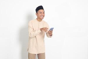 Portrait of young excited Asian muslim man in koko shirt with peci holding mobile phone with smiling expression on face. Social media and internet concept. Isolated image on white background photo