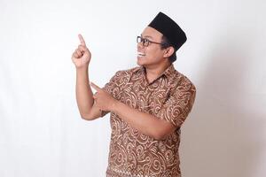 Portrait of excited Asian man wearing batik shirt and songkok smiling and looking at the camera pointing with two hands and fingers to the side. Isolated image on gray background photo