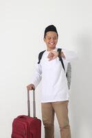 Portrait of displeased Asian muslim man standing over white background, holding the suitcase handle, paying attention to the time on watch preparing for the trip. Going home for Eid Mubarak. photo