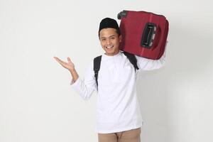 Portrait of excited Asian muslim man carrying suitcase pointing with hand to the side. Going home for Eid Mubarak. Isolated image on white background photo