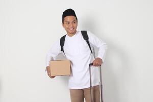 Portrait of excited Asian muslim man in koko shirt with peci carrying cardboard box and holding suitcase handle. Going home for Eid Mubarak. Isolated image on white background photo