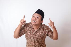 Portrait of excited Asian man wearing batik shirt and songkok smiling and looking at the camera pointing with two hands and fingers to up. Isolated image on gray background photo
