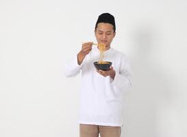 Portrait of attractive Asian muslim man eating tasteful instant noodles with chopsticks served on bowl. Iftar and pre dawn meal concept. Isolated image on white background photo