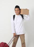 Portrait of cheerful Asian muslim man carrying suitcase and cardboard box while preparing for the flight. Going home for Eid Mubarak. Isolated image on white background photo