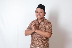 Portrait of smiling Asian man wearing batik shirt and songkok showing apologize and welcome hand gesture. Ramadan advertising concept. Isolated image on gray background photo