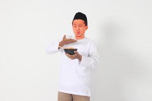 Portrait of shocked Asian muslim man eating tasty and spicy noodle and turning his face become red. Iftar and pre dawn meal concept. Isolated image on white background photo