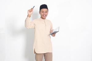 Portrait of attractive Asian muslim man in koko shirt with peci reading a book, telling that he has an idea while pointing finger and pen. Isolated image on white background photo