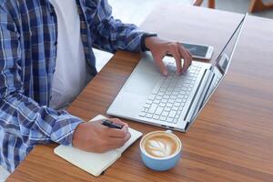 cerca arriba de masculino mano escritura en Nota libro con bolígrafo, demostración blanco pantalla de ordenador portátil y teléfono inteligente trabajando en café concepto con un taza de café foto