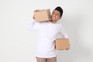 Portrait of tired Asian muslim man in koko shirt with peci having difficulty carrying cardboard. Going home for Eid Mubarak. Isolated image on white background photo