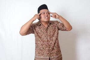 Portrait of suffering Asian man wearing batik shirt and songkok having headache while touching his forehead area. Isolated image on gray background photo