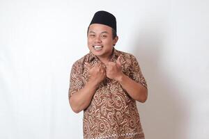 Portrait of excited Asian man wearing batik shirt and songkok smiling and looking at camera, making thumbs up hand gesture. Isolated image on gray background photo