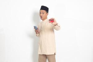 Portrait of attractive Asian muslim man in koko shirt with skullcap holding a mobile phone and presenting credit card. Isolated image on white background photo