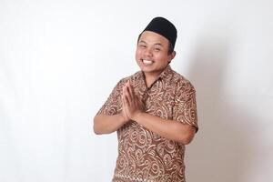 Portrait of smiling Asian man wearing batik shirt and songkok showing apologize and welcome hand gesture. Ramadan advertising concept. Isolated image on gray background photo