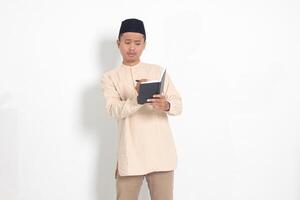 Portrait of excited muslim man in koko shirt with peci writing on his notebook. Happy Asian guy taking notes. Isolated image on white background photo