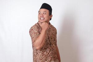 Portrait of excited Asian man wearing batik shirt and songkok smiling and looking at the camera pointing with two hands and fingers to the side. Isolated image on gray background photo