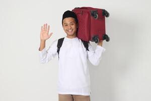 Portrait of attractive Asian muslim man carrying suitcase while saying hi to greet someone. Going home for Eid Mubarak. Isolated image on white background photo