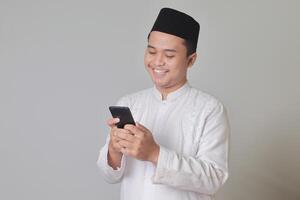 Portrait of young Asian muslim man holding mobile phone with smiling expression on face. Isolated image on white background photo