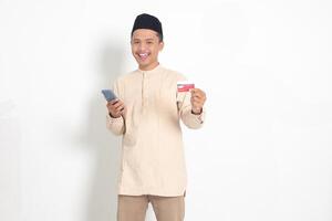 Portrait of attractive Asian muslim man in koko shirt with skullcap holding a mobile phone and presenting credit card. Isolated image on white background photo