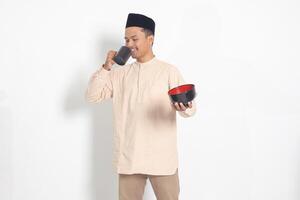 Portrait of excited Asian muslim man in koko shirt with skullcap holding a mug and empty bowl. Bowl template for food brand. Isolated image on white background photo