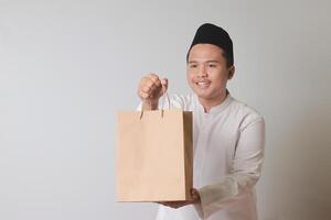 retrato de asiático musulmán hombre en blanco koko camisa con casquete dando arte papel compras bolso con regalo adentro. aislado imagen en gris antecedentes foto