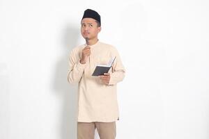 Portrait of confused Asian muslim man in koko shirt with peci reading a book, thinking about an idea with hand holding pen on chin, looking away. Isolated image on white background photo