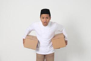 Portrait of surprised Asian muslim man in koko shirt with peci carrying cardboard box and making shocked expression. Going home for Eid Mubarak. Isolated image on white background photo