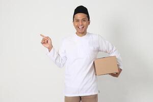 Portrait of attractive Asian muslim man in koko shirt with peci pointing to the side while carrying cardboard box. Going home for Eid Mubarak. Isolated image on white background photo