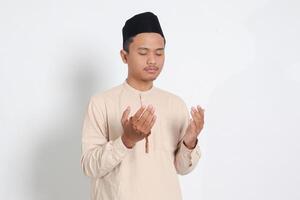 Portrait of religious Asian muslim man in koko shirt with skullcap praying earnestly with his hands raised, holding islamic beads. Devout faith concept. Isolated image on white background photo