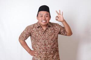 Portrait of excited Asian man wearing batik shirt and songkok showing ok hand gesture and smiling looking at camera. Isolated image on gray background photo