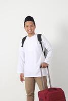 Portrait of attractive Asian muslim man standing against white background while holding the suitcase handle, preparing for the trip. Going home for Eid Mubarak. photo