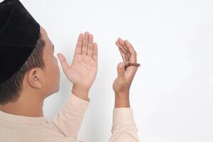 Back view portrait of religious Asian muslim man in koko shirt with skullcap praying earnestly with his hands raised, holding islamic beads. Devout faith concept. Isolated image on white background photo